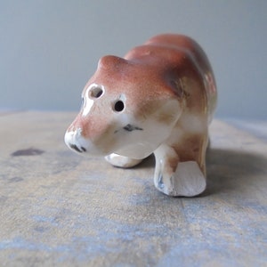 May include: A small, ceramic bear figurine. The bear is primarily brown with white accents on the face and feet. It has two eye holes and a small black nose. The figurine is standing on all fours.