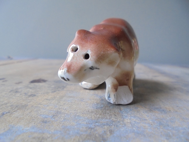 May include: A small, ceramic bear figurine. The bear is primarily brown with white accents on the face and feet. It has two eye holes and a small black nose. The figurine is standing on all fours.