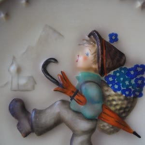 May include: A decorative ceramic plate with a figurine of a child carrying an umbrella and a basket of blue flowers. The child wears a brown hat, orange gloves, and a teal jacket. The plate has a white background with embossed details.