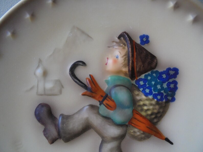 May include: A decorative ceramic plate with a figurine of a child carrying an umbrella and a basket of blue flowers. The child wears a brown hat, orange gloves, and a teal jacket. The plate has a white background with embossed details.