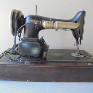 Op de afbeelding: Een vintage zwarte naaimachine met een houten basis en een metalen lamp. De machine heeft een voetpedaal en een naald.