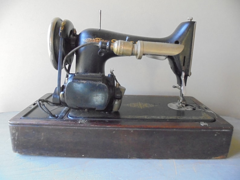Op de afbeelding: Een vintage zwarte naaimachine met een houten basis en een metalen lamp. De machine heeft een voetpedaal en een naald.