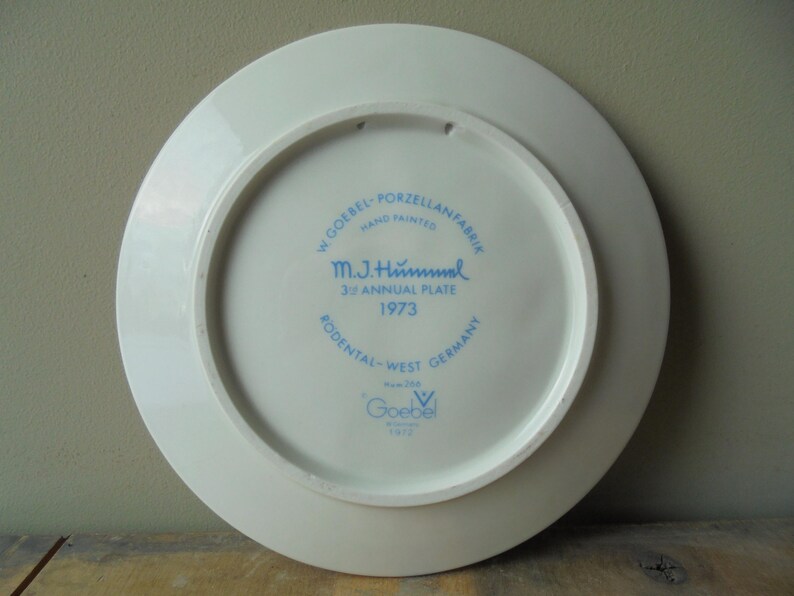 May include: A white porcelain plate with blue text and a circular design. The text includes "W. Goebel - Porzellanfabrik", "M.J. Hummel 39 Annual Plate 1973", and "Rodental-West Germany". The plate is hand-painted.