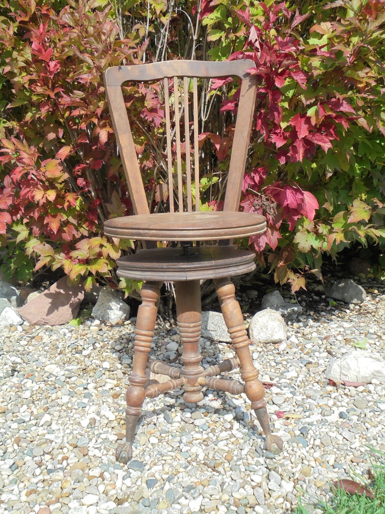 Pu&ograve; includere: Una sedia in legno vintage con schienale a fuso e seduta rotonda. La sedia ha una base girevole e quattro gambe con piedi a zampa. La sedia &egrave; marrone e ha una finitura usurata. La sedia &egrave; seduta su un letto di ghiaia.