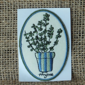 May include: A white oval embroidered patch with a green thyme plant in a blue and green striped pot. The text "Thyme" is embroidered in blue below the pot.