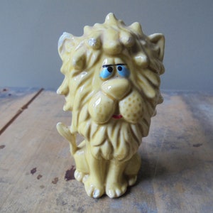May include: A vintage ceramic lion figurine in a seated position. The lion has a yellow body, a detailed mane, and a red mouth. Its eyes are blue, and it has a slightly distressed appearance.