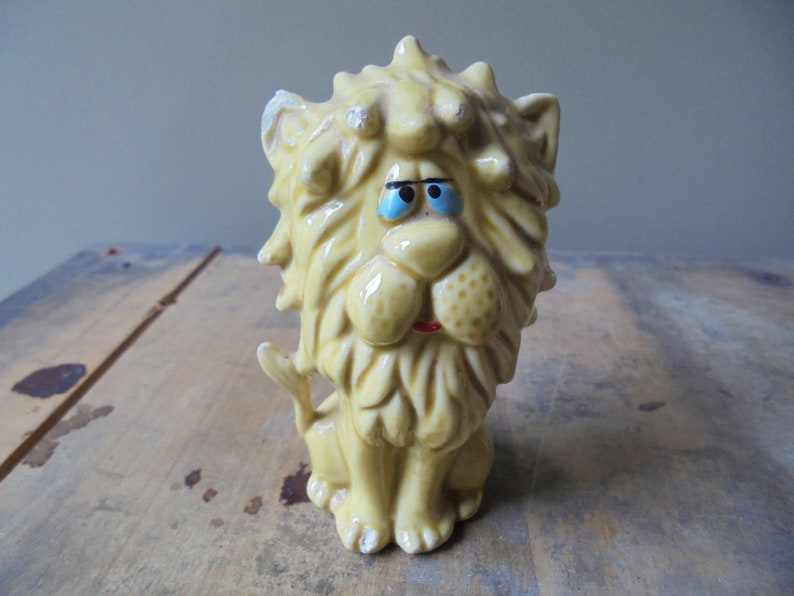 May include: A vintage ceramic lion figurine in a seated position. The lion has a yellow body, a detailed mane, and a red mouth. Its eyes are blue, and it has a slightly distressed appearance.