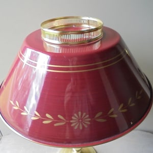 May include: A vintage red lamp shade with gold accents. The shade has a floral and leaf design. The base is gold-colored metal. The lamp shade is a cone shape with a gold ring at the top.