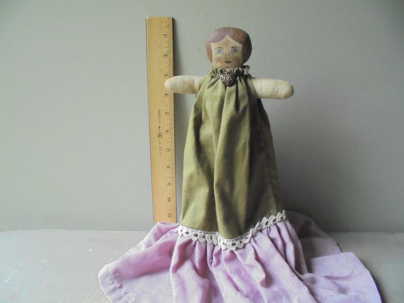 K&ouml;nnte beinhalten: Eine Vintage-Stoffpuppe mit bemaltem Gesicht und braunen Haaren. Die Puppe tr&auml;gt ein gr&uuml;nes Kleid mit wei&szlig;er Spitzenborte und einen rosa Rock.