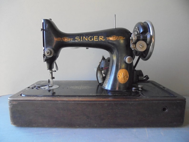 Op de afbeelding: Een vintage zwarte Singer naaimachine met een houten basis. De machine heeft een gouden naamplaat met het woord "SINGER" erop.