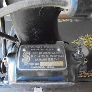 Op de afbeelding: Close-up van een vintage naaimachine motor met een zilveren label met de tekst "Sewing Moto Patented U.S.A. 100-110 Volts 0.6 Amps D.C. & 25 to 75 Cycles Catalog B.U.7-E Motor No. 4730840 Simanco, U.S.A."