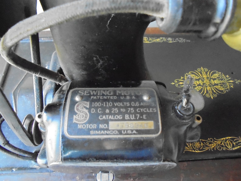 Op de afbeelding: Close-up van een vintage naaimachine motor met een zilveren label met de tekst "Sewing Moto Patented U.S.A. 100-110 Volts 0.6 Amps D.C. & 25 to 75 Cycles Catalog B.U.7-E Motor No. 4730840 Simanco, U.S.A."