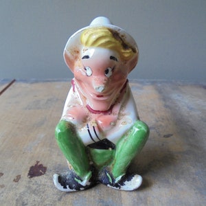 May include: A vintage ceramic figurine of a seated elf-like character. The figurine has a white hat, blonde hair, and a pink face. It wears a white shirt, green pants, and black shoes. The figurine is sitting with its legs crossed.