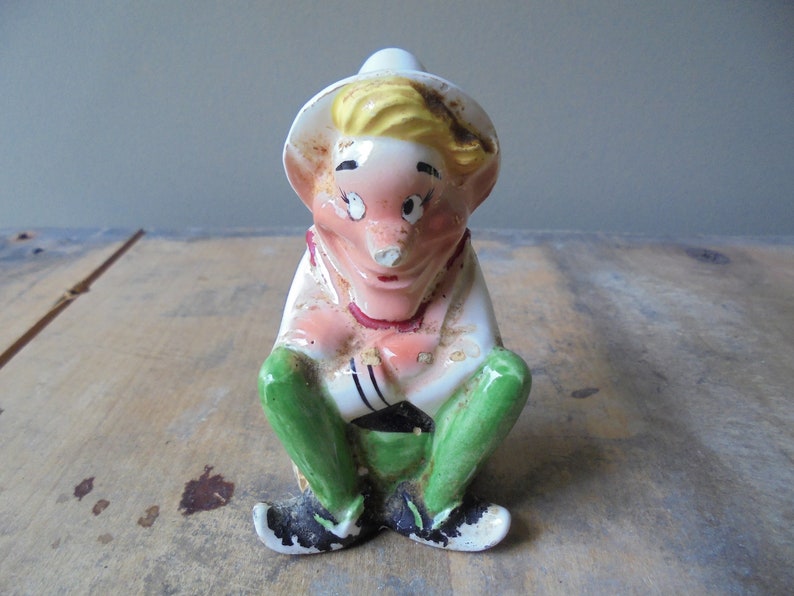 May include: A vintage ceramic figurine of a seated elf-like character. The figurine has a white hat, blonde hair, and a pink face. It wears a white shirt, green pants, and black shoes. The figurine is sitting with its legs crossed.