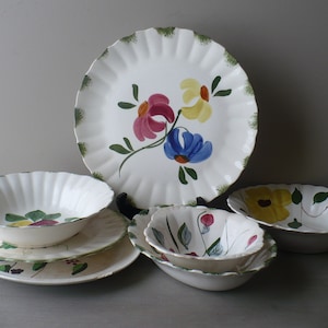 May include: A collection of white ceramic tableware with scalloped edges and hand-painted floral designs. The set includes plates and bowls, featuring colorful flowers in shades of pink, yellow, and blue, with green leaves.