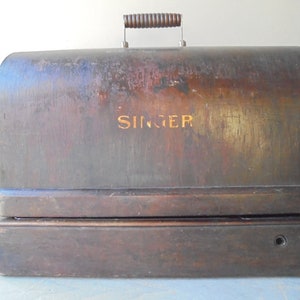 Op de afbeelding: Een vintage houten Singer naaimachine koffer met een versleten bruine afwerking en een metalen handvat. De koffer heeft een gouden inscriptie met de tekst "SINGER".
