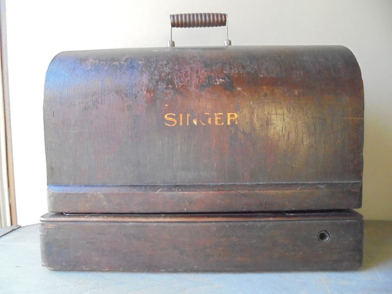 Op de afbeelding: Een vintage houten Singer naaimachine koffer met een versleten bruine afwerking en een metalen handvat. De koffer heeft een gouden inscriptie met de tekst "SINGER".