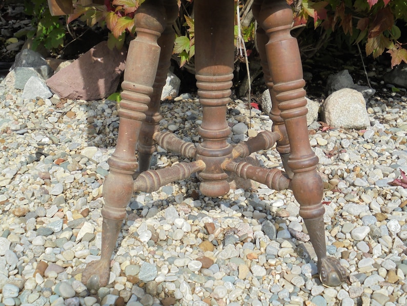 Pu&ograve; includere: Uno sgabello di legno con piano rotondo e tre gambe. Le gambe sono tornite e hanno un design decorativo. Lo sgabello &egrave; seduto su un letto di ghiaia.