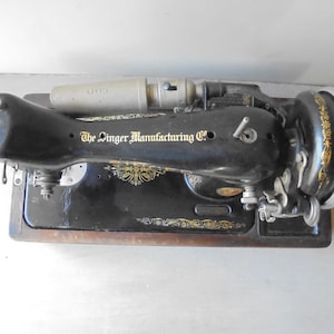 Op de afbeelding: Zwarte vintage Singer naaimachine met een houten basis en een metalen voetpedaal. De machine heeft een gouden logo met de tekst "The Singer Manufacturing Co."