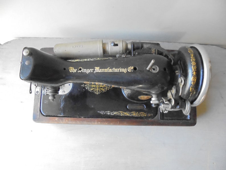 Op de afbeelding: Zwarte vintage Singer naaimachine met een houten basis en een metalen voetpedaal. De machine heeft een gouden logo met de tekst "The Singer Manufacturing Co."
