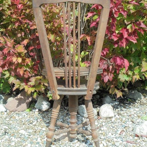 Pu&ograve; includere: Una sedia girevole in legno vintage con schienale a doghe e base a forma di stella. La sedia &egrave; marrone e ha una finitura usurata. &Egrave; seduta su un letto di ghiaia.