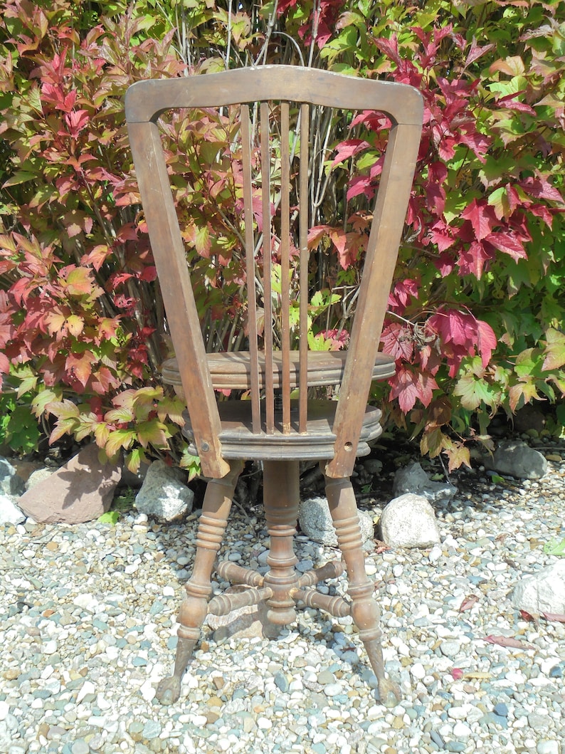 Pu&ograve; includere: Una sedia girevole in legno vintage con schienale a doghe e base a forma di stella. La sedia &egrave; marrone e ha una finitura usurata. &Egrave; seduta su un letto di ghiaia.