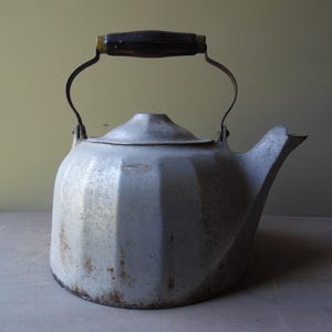 May include: A vintage-style metal tea kettle with a light gray, fluted body and a wide, integrated spout. The kettle features a curved metal handle attached to a dark brown, rectangular grip at the top, showing signs of wear and oxidation.