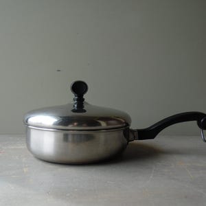 May include: Stainless steel saucepan with a lid and a black handle. The pan has a rounded body and a lid with a black knob. The handle has a loop at the end. The pan is sitting on a light gray surface.