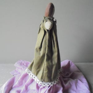 K&ouml;nnte beinhalten: Eine handgemachte Puppe, die ein gr&uuml;nes Kleid mit wei&szlig;er Spitzenborte tr&auml;gt. Die Puppe sitzt auf einem violetten Stoff.