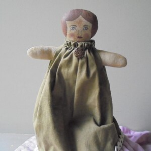 K&ouml;nnte beinhalten: Eine handgemachte Stoffpuppe mit einem braunen und gr&uuml;nen Kleid und einem braunen Knopf. Die Puppe hat gemalte braune Haare, blaue Augen und ein gemaltes Gesicht.