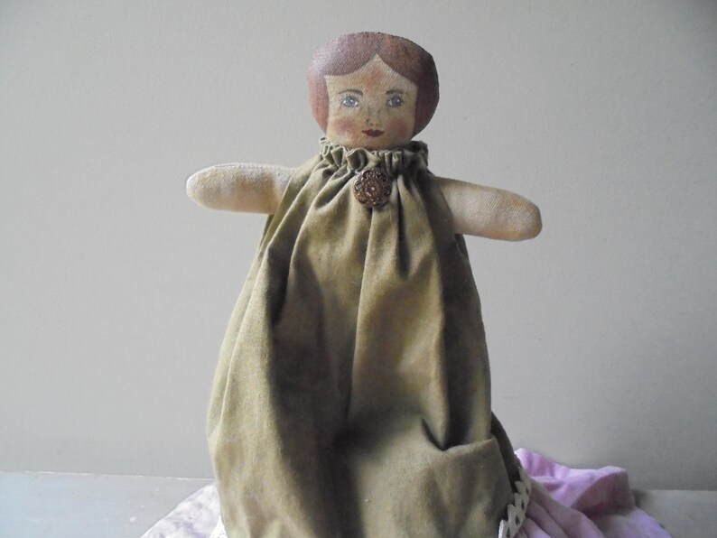 K&ouml;nnte beinhalten: Eine handgemachte Stoffpuppe mit einem braunen und gr&uuml;nen Kleid und einem braunen Knopf. Die Puppe hat gemalte braune Haare, blaue Augen und ein gemaltes Gesicht.