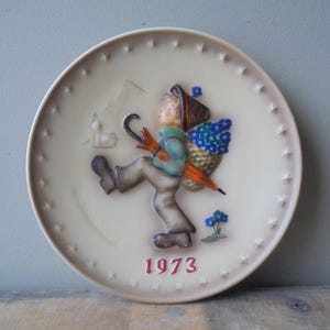 May include: A cream-coloured decorative plate with a relief design. It depicts a figure carrying a basket of blue flowers and an orange umbrella. The plate has a star-studded border and the year "1973" in red.