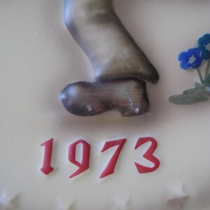 May include: A cream-coloured decorative piece featuring a sculpted figure's leg and shoe, with the year "1973" in red script. Blue flowers with green leaves are also present. The shoe is brown and the leg is a muted grey.