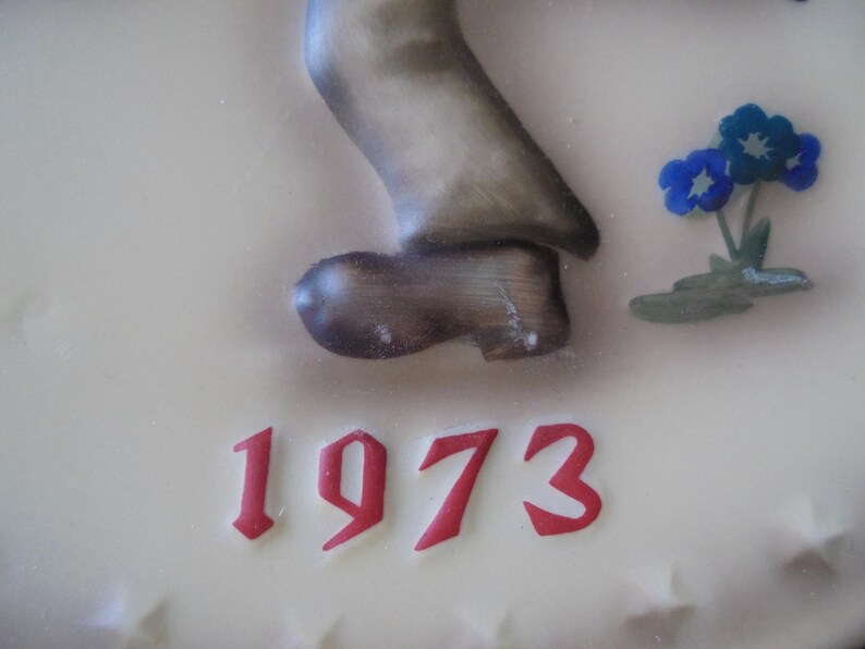 May include: A cream-coloured decorative piece featuring a sculpted figure's leg and shoe, with the year "1973" in red script. Blue flowers with green leaves are also present. The shoe is brown and the leg is a muted grey.