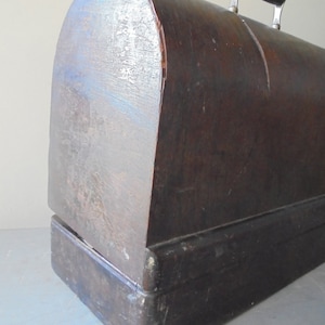 Op de afbeelding: Een vintage houten naaimachinekoffer met een donkerbruine afwerking en een metalen handvat. De koffer heeft een afgeronde bovenkant en een rechthoekige basis.