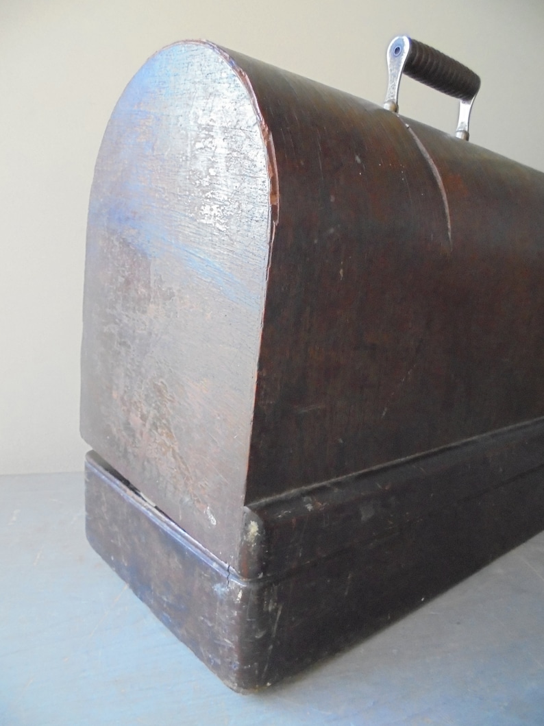 Op de afbeelding: Een vintage houten naaimachinekoffer met een donkerbruine afwerking en een metalen handvat. De koffer heeft een afgeronde bovenkant en een rechthoekige basis.