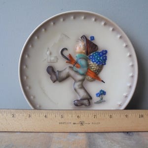 May include: A decorative ceramic plate with a relief of a child walking with an umbrella and a basket of blue flowers. The plate has a cream background with star-shaped accents around the rim. A wooden ruler is placed below the plate.