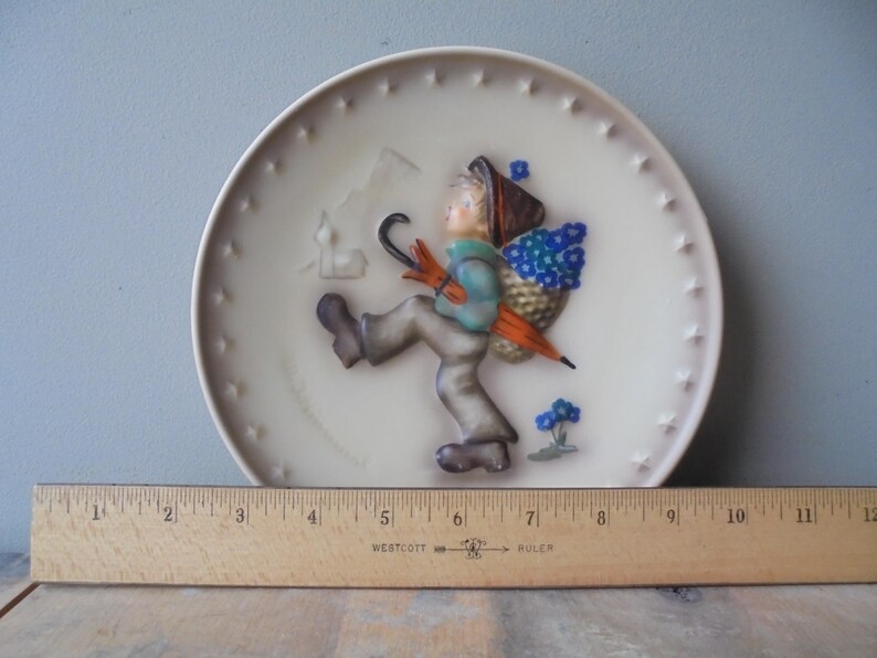 May include: A decorative ceramic plate with a relief of a child walking with an umbrella and a basket of blue flowers. The plate has a cream background with star-shaped accents around the rim. A wooden ruler is placed below the plate.