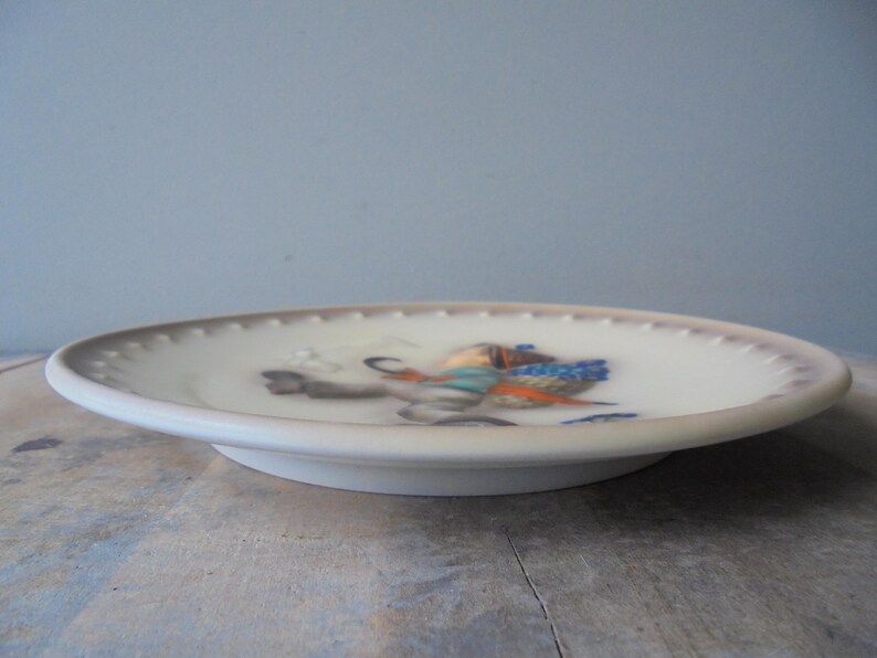 May include: A white ceramic plate with a detailed illustration of a bird in flight, featuring blue, orange, and grey hues. The plate has a slightly raised rim with a dotted pattern and a diameter of approximately 15 cm.