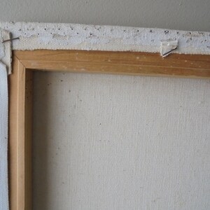 May include: Close-up of the top edge of a canvas painting. The canvas is stretched over a wooden frame and secured with staples. The canvas is white and the frame is a light brown wood.