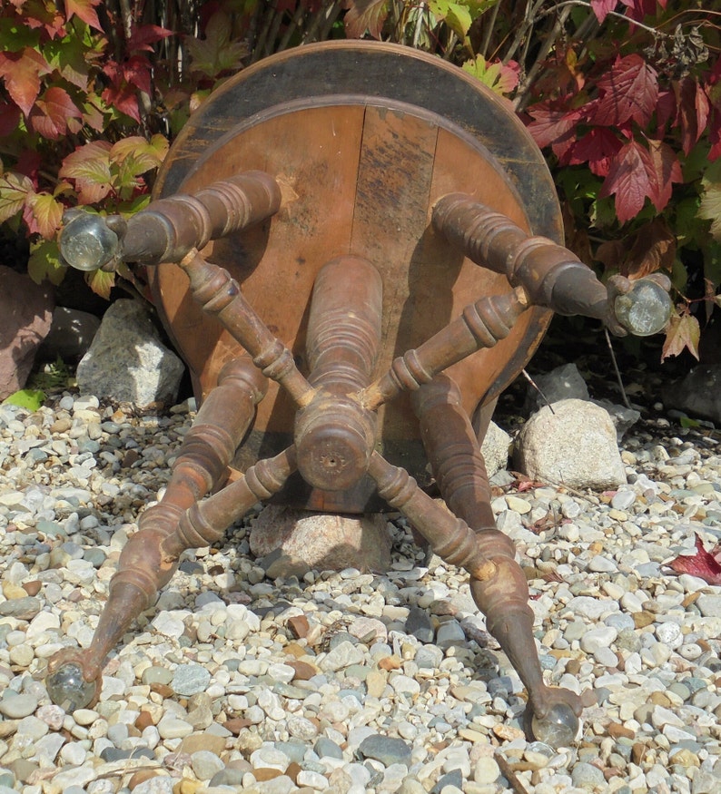 Pu&ograve; includere: Uno sgabello in legno antico con piano rotondo e quattro gambe. Lo sgabello &egrave; dipinto di marrone e ha un bordo nero attorno al piano. Le gambe sono tornite e presentano dettagli decorativi. Lo sgabello &egrave; seduto su un letto di ghiaia.
