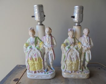 Può includere: Coppia di lampade da tavolo in porcellana con una coppia in abiti del XVIII secolo. Le lampade sono bianche con dettagli dorati e hanno un'estetica vintage.