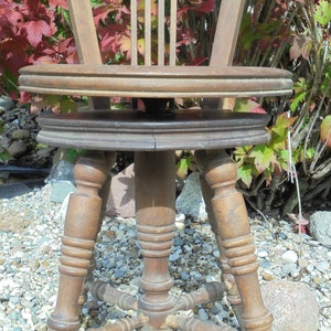 Pu&ograve; includere: Una sedia girevole in legno con un sedile rotondo e gambe tornite. La sedia &egrave; in un giardino con ghiaia sul terreno.