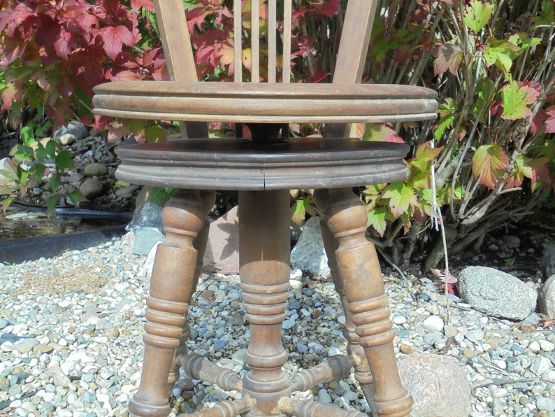 Pu&ograve; includere: Una sedia girevole in legno con un sedile rotondo e gambe tornite. La sedia &egrave; in un giardino con ghiaia sul terreno.