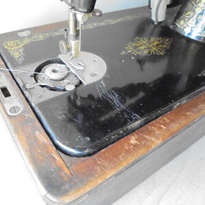 Op de afbeelding: Een vintage zwarte naaimachine met gouden accenten en een houten basis. De machine heeft een spoelwinder en een naaldinvoer.