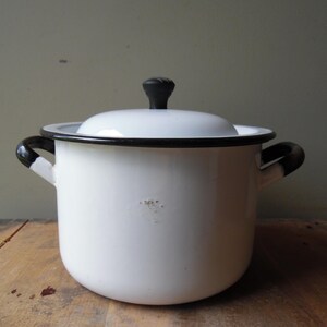 May include: A white enamel pot with a matching lid, featuring a black rim and two black side handles. The pot has a cylindrical body with a small area of surface wear on the front. The lid is topped with a black knob for easy handling.