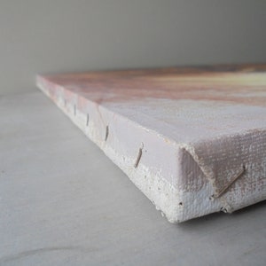 May include: Close-up of the edge of a canvas painting. The canvas is stretched over a wooden frame and secured with staples. The canvas is a light beige color and the staples are silver.