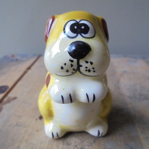 May include: A ceramic dog figurine with a yellow body, brown ears, and a black nose. The dog has large eyes and is sitting upright. The figurine is painted with black details and has a glossy finish. The dog is a decorative item.