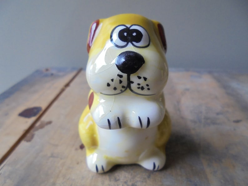 May include: A ceramic dog figurine with a yellow body, brown ears, and a black nose. The dog has large eyes and is sitting upright. The figurine is painted with black details and has a glossy finish. The dog is a decorative item.
