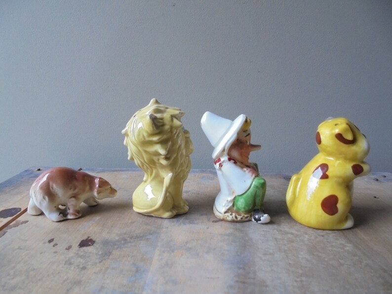 May include: A collection of four ceramic figurines: a brown bear, a yellow lion, a gnome, and a yellow dog with brown spots. The figurines are arranged in a row, each with unique details and colors.